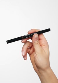 Matita eyeliner nera con etichetta "BOBBI BROWN" e testo "Rich Brown", tenuta in mano, con un design liscio ed elegante.