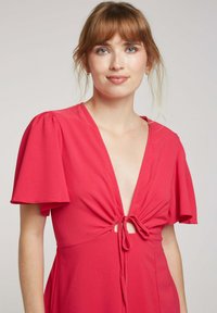 Robe rouge avec un décolleté en V profond, des manches bouffantes courtes et un détail noué à la taille. Fabriquée dans un tissu léger et texturé.