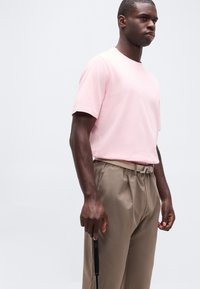 Άνδρας που φοράει ανοιχτό ροζ μπλουζάκι και μπεζ παντελόνι κρατώντας ένα γκολφ κλαμπ μπροστά σε απλό ανοιχτό φόντο.