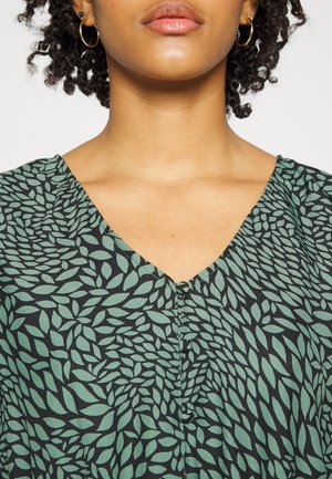 Woman with gold hoop earrings wearing a black V-neck top with a green leafy pattern.