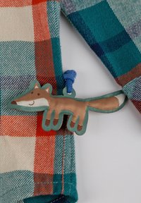 Camicia a quadri con colori rosso, teal e crema. Presenta un'etichetta in tessuto a forma di volpe con un'asola blu. La volpe ha un volto sorridente e una coda.