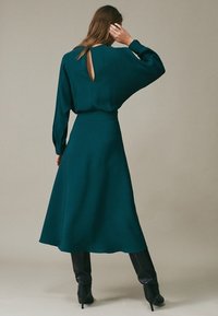 Donna con un abito lungo maniche teal e un dettaglio a chiave sul retro, abbinato a stivali neri alti fino al ginocchio con tacco, in piedi contro uno sfondo semplice.