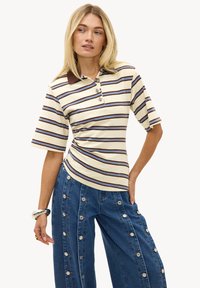 Femme blonde portant un polo à rayures crème et marron associé à un jean large bleu avec plusieurs boutons sur les côtés.
