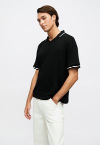 Νέος άνδρας με καστανά μαλλιά που φοράει μαύρο πλεκτό πόλο με κοντά μανίκια και λευκή επένδυση και λευκό παντελόνι, με τα χέρια στις τσέπες, ουδέτερο φόντο.