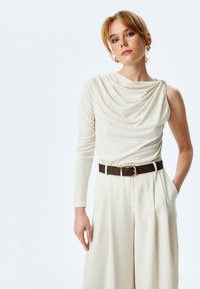 Haut créme drapé avec une manche longue, associé à un pantalon créme taille haute à plis. Ceinture marron foncé avec une boucle dorée en accent.