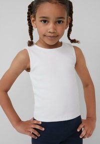 Jong meisje met een gevlochten haar, gekleed in een mouwloze witte top en donkere broek, staand met handen op de heupen tegen een effen achtergrond.