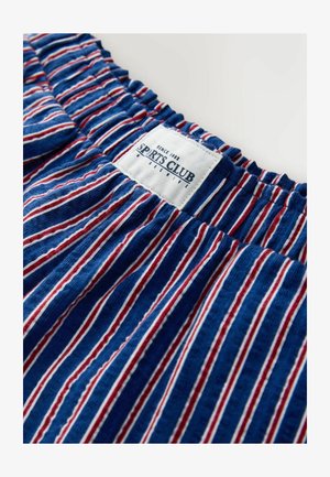 Dryžuotas audinys mėlynos, raudonos ir baltos spalvų, su susmulkinta elastine iškirpte ir baltu etikete, kurioje parašyta „SPORTS CLUB“. Tekstūra atrodo minkšta.