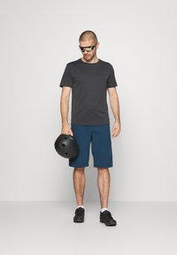Dunkelgraues T-Shirt mit kurzen Ärmeln, blaue Shorts, schwarzen Helm in der Hand, schwarze Schuhe und Sonnenbrille mit gelben Akzenten. Schlichtes und funktionales Design.