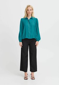 Blusa teal a maniche lunghe con bottoni frontali e polsini elasticizzati, abbinata a pantaloni neri a gamba larga e sandali neri con cinturino.