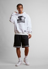 Hvid hættejakke med sort logo, sorte shorts med hvid stribe, hvide sneakers og ankelstrømper. Modellen står foran en neutral baggrund.