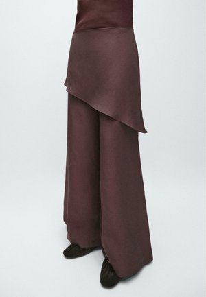 Brune bukser med vide ben og asymmetrisk lagdelt stofdetalje på den øverste lårdel, kombineret med sorte sko med blød tekstur.