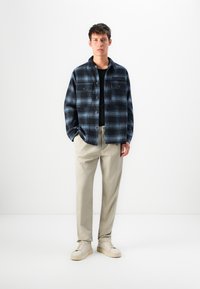 Giovane uomo che indossa una camicia a quadri blu e nera sopra una maglietta nera, pantaloni beige e sneakers beige, in piedi di fronte a uno sfondo bianco.