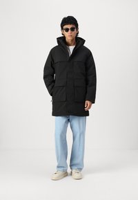 Black waterproof parka with a hood, featuring two front pockets and a straight cut. Worn over light blue trousers and beige sneakers.