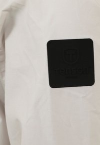 Lichtgekleurde stof met een gladde textuur heeft een zwart rubberen logopatch met de tekst "tenson" erop geperst.