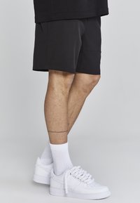 Black shorts with a relaxed fit, cotton fabric, knee-length, paired with white sneakers and ribbed white socks. Features a subtle seam detail.
