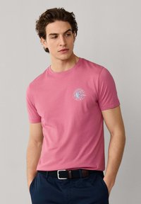 Giovane uomo con capelli ricci scuri che indossa una t-shirt rosa aderente con un piccolo logo bianco sul petto e pantaloni blu navy con cintura.
