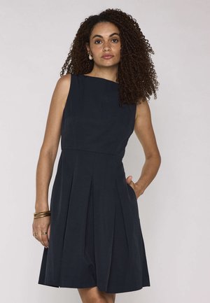 Vrouw met krullend haar die een mouwloze marineblauwe jurk met plooien draagt, gouden armbanden, ringen en pareloorbellen, poserend met één hand in de zak.