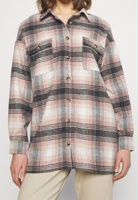 Plaid flannel shirt in pink, black, and white. Features two chest pockets, buttons, and a relaxed fit with long sleeves. Soft texture.