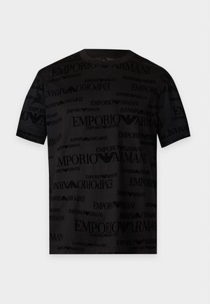 Jeune homme debout, portant un t-shirt noir avec le logo Emporio Armani, un jean bleu clair retroussé et des baskets blanches sur fond blanc.