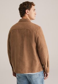 Bruine suede jas met een kraag en knoopmanchetten, met een gladde structuur en een relaxed fit, gecombineerd met lichtblauwe spijkerbroek.