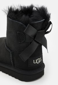 Bottine en suède noire avec doublure en fourrure noire duveteuse, un nœud en ruban noir décoratif sur le côté et un logo UGG blanc à l'arrière.