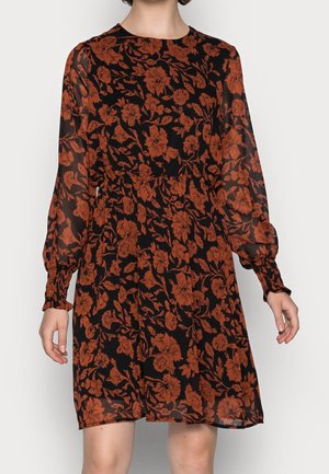 Femme portant une robe noire longueur genou avec manches longues et un motif floral rouille sur toute la surface, debout devant un fond uni.