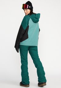 Veste de ski teal et noire, en tissu imperméable, avec une capuche. Accompagnée d'un pantalon teal assorti et de bottes noires. Les accessoires comprennent un bonnet noir et des lunettes de ski miroitées.
