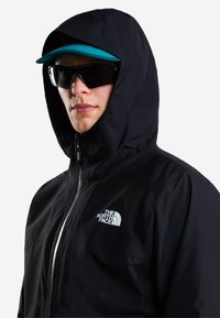 Άτομο που φοράει μαύρο μπουφάν North Face με κουκούλα, τυρκουάζ καπέλο και μεγάλα μαύρα γυαλιά ηλίου πάνω σε λευκό φόντο.