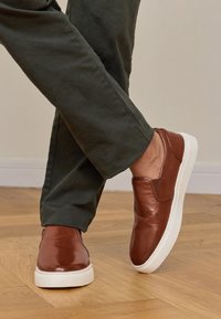 Person, der braune Leder-Einschubschuhe mit weißen Sohlen trägt und in einer dunklen grünen Hose auf einem Holzfußboden steht.