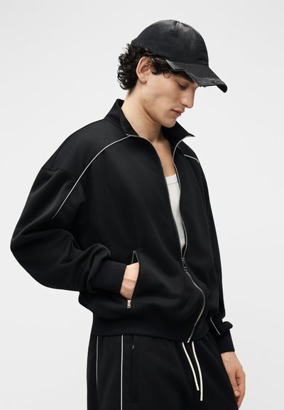 Νεαρός άνδρας με μαύρο μπουφάν με φερμουάρ και παντελόνι που ταιριάζει με λευκές ρίγες, φορώντας μαύρο καπέλο μπέιζμπολ, κοιτάζοντας προς τα κάτω με το ένα χέρι στην τσέπη.