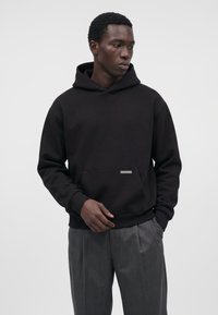 Homme portant un sweat à capuche noir et un pantalon gris plissé, debout avec une main dans la poche devant un fond uni.