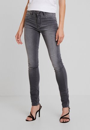 Kvinde iført tætsiddende grå jeans og sorte højhælede sandaler, stående mod en ensartet lysegrå baggrund.