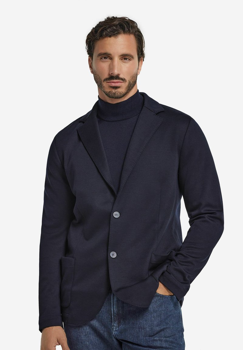 Mężczyzna z krótkimi, kręconymi włosami i brodą, ubrany w granatową marynarkę na sweter z golfem oraz niebieskie dżinsy, z jedną ręką w kieszeni.