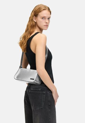 Metallische silberne Handtasche mit strukturiertem Design, glatter Textur und schwarzem Besatz, lässig über einem schwarzen Tanktop getragen.