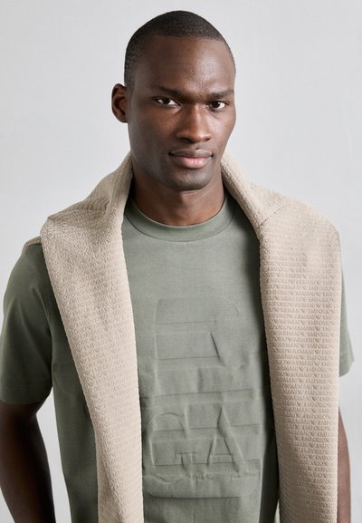 T-shirt in cotone verde oliva con logo testurizzato, abbinato a una sciarpa in pile beige e motivo, poggiata sulle spalle.
