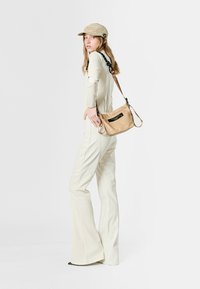 Gorra beige, top de manga larga crema y pantalones acampanados. Sostiene un bolso pequeño marrón claro con una etiqueta negra, que presenta una textura suave. Diseño minimalista.