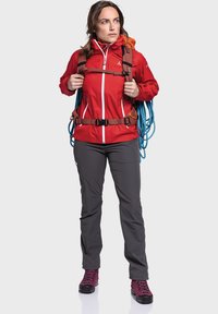 Red waterproof jacket with white zippers, gray outdoor pants, and burgundy hiking boots. Model carries a brown harness and blue rope.