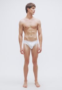 Ανδρικά λευκά σλιπ με απλό σχέδιο, με λαστιχένια μέση που φέρει λογότυπο. Το ύφασμα φαίνεται λείο και ελαφρύ.