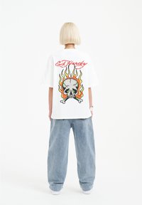 Bílá bavlněná tričko s velkým potiskem lebky a plamenů na zadní straně. Vyznačuje se textem "Ed Hardy" v červené barvě nad designem. Nosené s volnými modrými džíny.