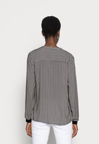 Blusa de mangas longas preta e branca com padrão, punhos elásticos, decote redondo e corte ligeiramente solto, combinada com calças brancas.