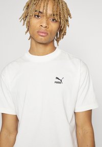 Bílý bavlněný T-shirt s černým logem Puma na levé hrudi, kulatým výstřihem a krátkými rukávy. Hladká textura, volný střih.