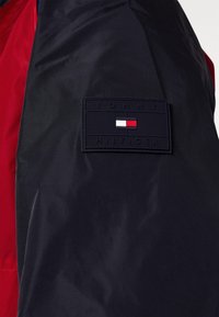 Giacca a blocchi di colore blu navy e rossa con un tessuto texturizzato che presenta una toppa rettangolare blu navy. Sulla toppa è riportato "TOMMY HILFIGER" e un logo in rosso, bianco e blu.