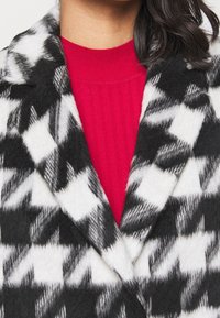 Sort og hvid houndstooth-mønstret uldfrakke med dyb V-udskæring og struktureret krave, lagt over en rød ribbet sweater.