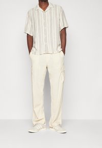 Kortärmad, kragad skjorta med vertikala ränder i beige och blått. Matchas med löst sittande, krämfärgade cargobyxor och ljusa sneakers.