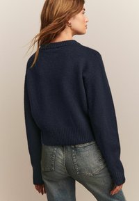 Maglione corto blu navy realizzato in tessuto a maglia testurizzato con polsini e orlo a coste. Abbinato a jeans blu sbiaditi, visto da dietro.