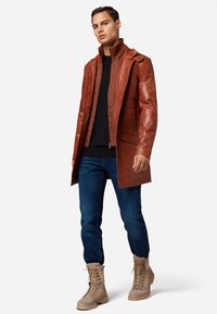 Bruine leren jas met hoge kraag, gelaagd over een zwarte trui. Draag met blauwe jeans en tan kleurige veterschoenen. Geweven ontwerpdetails zichtbaar.