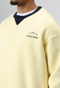Gele trui met een V-halsontwerp, voorzien van een zwarte geribbelde kraag en de tekst "LE-75005-DE STUDIO ABYSS" op de borst. Zachte textuur.