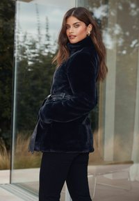 Mākslīgās kažokādas mētelis tumši zils ar augstu apkakli un jostu viduklī. Mīksta tekstūra ar plūksu apdari, apvienots ar melniem pieguļošiem biksēm.