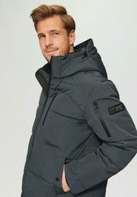 Padded szary kurtka w ciemnym kolorze z zapięciem na zamek, regulowanym kapturem i bocznymi kieszeniami. Posiada naszywkę z logo na rękawie oraz teksturę materiału.