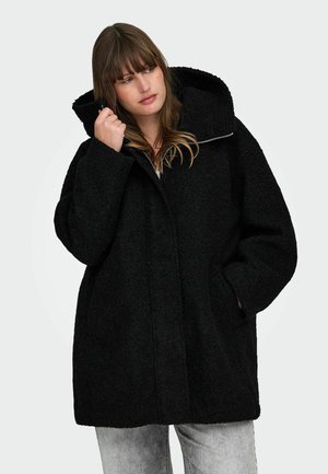 Must musta, tekstuuriga ülevõetud mantel kõrge kraega, lukuga kinnitamine ja suur kapuuts. Tal on küljetaskud ja lõdva, juhusliku lõikega.